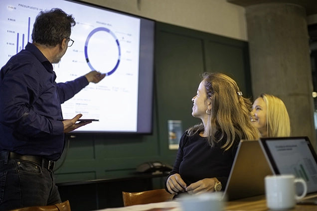 En mann gir økonomisk rådgivning, og presenterer data på en skjerm til to kvinner som sitter ved et bord med bærbare datamaskiner og kaffekopper i et møterom.
